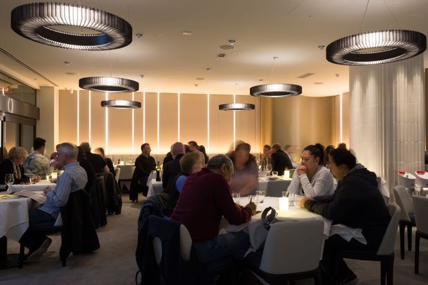 A busy contemporary restaurant dining room with numerous diners seated at tables. The space features warm beige and cream tones, vertical illuminated wall panels, a pale carpeted floor, and distinctive large circular pendant lights with dark ribbed detailing. White upholstered dining chairs and dark tables create a refined, softly lit atmosphere.