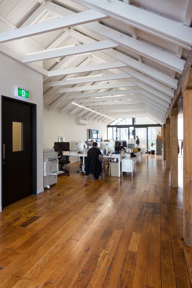 A bright open-plan office occupying a long adaptive-reuse-style interior, with rows of desks and computers, white-painted exposed timber roof trusses, high pitched ceilings, polished or worn warm timber floorboards, large glazed doors or windows at the far end, and substantial timber structural posts along the right side. A black emergency exit door and office equipment are visible on the left.