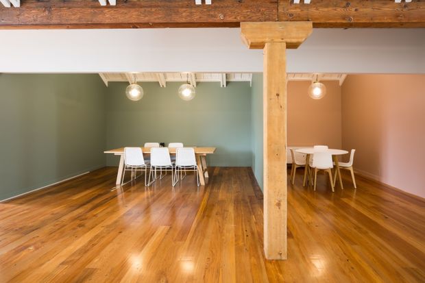 A spacious open-plan dining area with polished timber flooring, exposed timber beams and a prominent structural timber post. Two dining settings with white chairs are arranged against contrasting muted green and blush-pink walls. The ceiling features exposed painted joists and simple globe pendant lights, creating a warm, characterful interior.