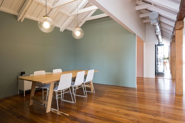 Open-plan dining area in a converted industrial-style interior, featuring a timber dining table with white chairs, polished timber flooring, a large muted green feature wall, exposed white pitched roof structure, hanging globe lights, and visible ductwork along the corridor.
