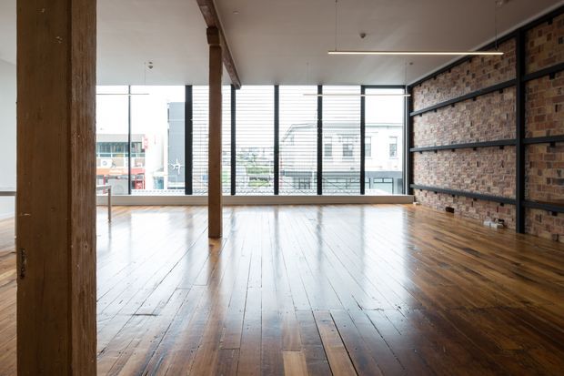 An expansive open-plan interior with polished dark timber floorboards, exposed timber posts and beams, and a full-height exposed brick feature wall crossed by dark steel framing and shelving. Large industrial-style windows provide abundant daylight and urban views. Minimal furnishings emphasize the loft-like character and generous floor area.