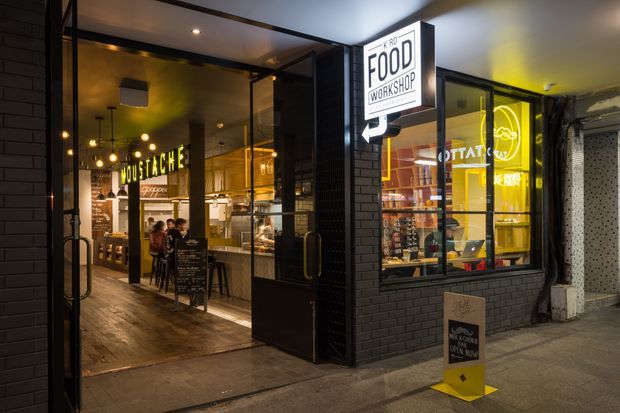 Night-time street-level view of a contemporary food workshop and restaurant frontage. The image shows a black-painted brick facade, large glazed shopfront windows and an open entry, with warm interior lighting, pendant lights, timber-look flooring, dining seating, food-service counters and prominent food-related signage. The scene is primarily an urban commercial hospitality venue viewed from outside.
