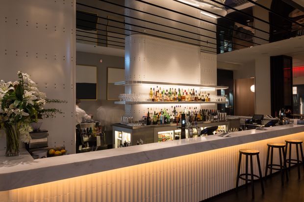 A sophisticated contemporary bar interior with a long pale stone bar counter, integrated warm underlighting, vertically ribbed bar-front cladding, and black bar stools. Behind the counter are illuminated shelves displaying bottles and glassware, with a large sculptural metallic canopy and perforated wall panels above. The space has a refined, atmospheric hospitality design with dark ceilings and dramatic accent lighting.