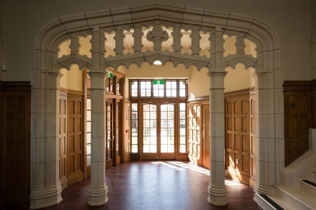 A grand historic interior entrance hall with ornate carved stone arches and columns framing a sunlit timber-panelled vestibule. Dark polished timber doors and wall panelling surround glazed double entry doors, while a staircase is partially visible at right. The space has strong Gothic Revival or Victorian character, with warm natural light and intricate architectural detailing.