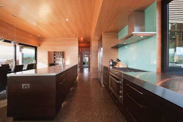 A contemporary galley-style kitchen with a large central island, dark timber cabinetry, stainless-steel appliances and benchtops, and a pale turquoise glass splashback. The room has extensive warm timber lining across the ceiling and walls, polished dark flooring, integrated lighting, and large windows offering views outside. A dining area is visible at the left, while the kitchen dominates the frame.