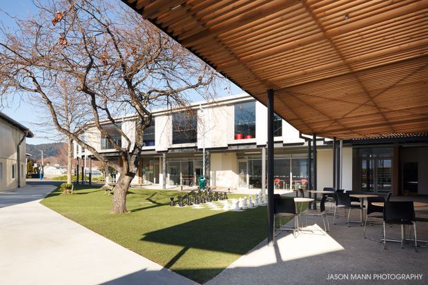 A bright contemporary courtyard or outdoor dining area beside a modern two-storey building. A timber slatted pergola with slim dark posts shelters tables and bar-height chairs, while a lawn, mature deciduous tree, oversized outdoor chess set, glazing and pale rendered facade define the shared courtyard. Mountainous terrain is visible in the distance.
