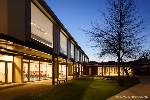 Architectural exterior photograph of a contemporary multi-wing building surrounding a landscaped courtyard at dusk. The two-storey facade features extensive floor-to-ceiling glazing, dark vertical framing, projecting upper-level volumes, and a covered ground-floor walkway. Warm interior lighting contrasts with the cool evening sky, while a lawn, mature deciduous tree, and paved path occupy the courtyard.