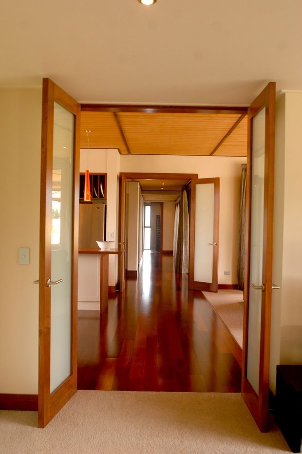 View through open timber-framed glazed double doors into a long, warm-toned interior corridor. The passage has highly polished dark timber flooring, cream walls, timber skirting and trims, and a distinctive timber-lined ceiling with exposed linear framing. Multiple open doors lead to adjoining rooms, with a partial kitchenette or vanity area visible on the left. The foreground room has a beige carpet and the image is primarily an architectural hallway view.