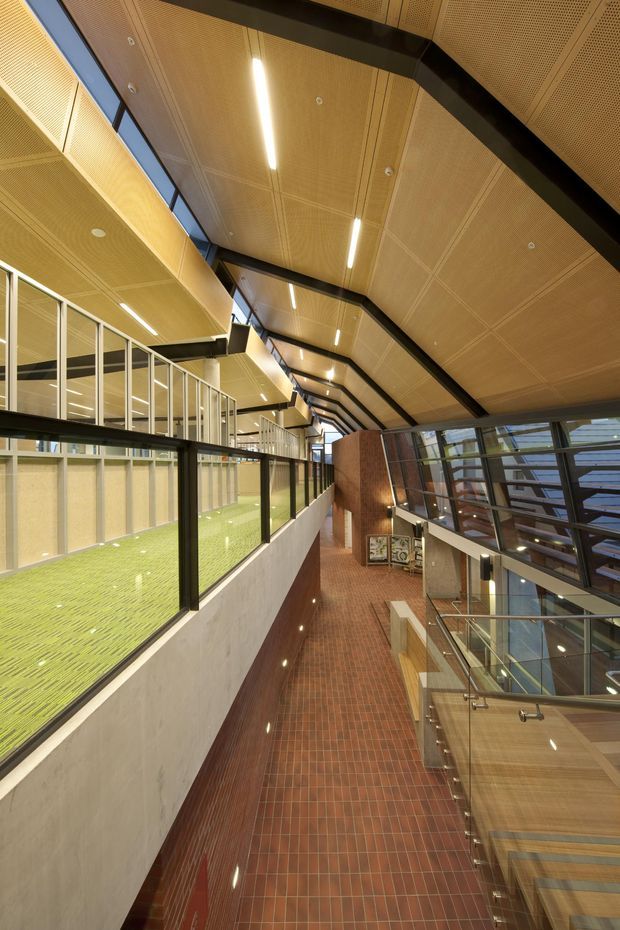 Interior view of a large contemporary institutional atrium or circulation space, featuring a high perforated timber-panel ceiling with integrated linear lighting, exposed black structural beams, extensive glazing, glass balustrades, and an elevated green-floored area overlooking a red brick-tiled passage. The space has a dramatic multi-level composition and abundant natural light.