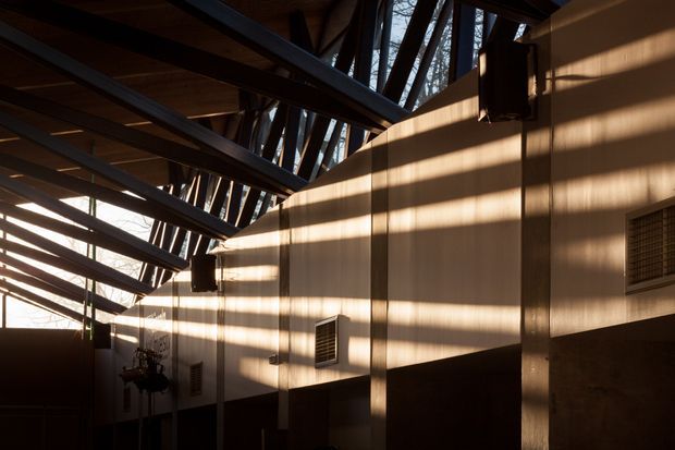 Interior architectural view of a long, industrial-style space with exposed dark steel roof trusses and timber roof lining. Warm sunlight passes through high-level glazing or openings, casting strong linear shadows across tan wall panels. Wall-mounted ventilation grilles, speakers, and exposed structural elements contribute to a utilitarian, dramatic atmosphere.