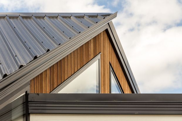 Close-up exterior architectural detail showing a steep gabled roof with corrugated metal roofing, warm vertical timber cladding, dark-framed triangular glazing, and a lower flat-roofed or projecting volume. The image emphasizes the junction of contrasting roof forms and facade materials against a cloudy sky.