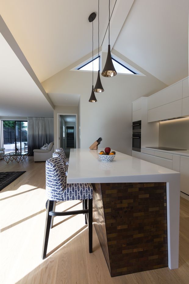 Bright open-plan kitchen and living area with a large white island, integrated white cabinetry, built-in ovens, and a dark tiled island base. Three black pendant lights hang beneath a high pitched ceiling with triangular clerestory windows. Light timber flooring, patterned bar stools, and a glimpse of a lounge area complete the contemporary interior.