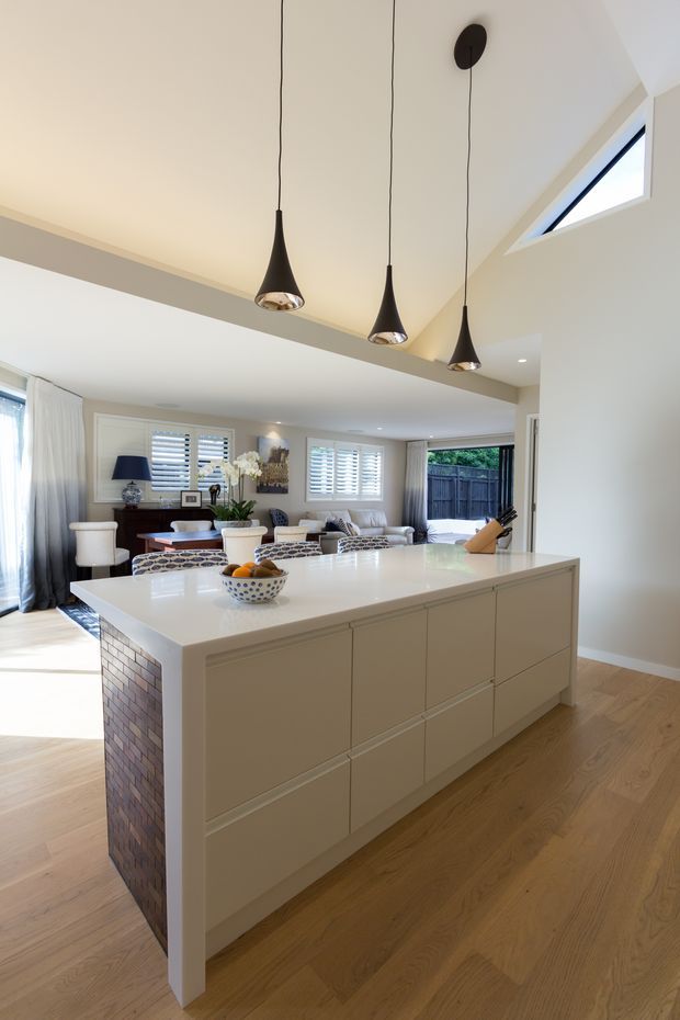 Bright open-plan kitchen and living area centered on a large white island with integrated panelled cabinetry and a pale stone benchtop. The island has a dark exposed brick end panel, while three black pendant lights hang beneath a high ceiling with a narrow skylight. Timber flooring, white walls, plantation shutters, upholstered seating, and a dining area create a refined contemporary residential interior.