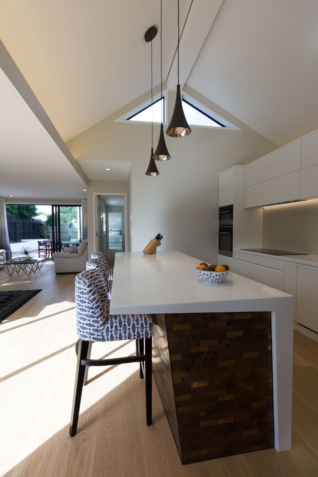 A bright, contemporary open-plan kitchen with a large white island, integrated white cabinetry, built-in appliances and under-cabinet lighting. The island has a dark brick-like tiled feature face and is paired with patterned upholstered bar stools. A double-height, pitched ceiling with triangular skylights and three suspended pendant lights forms a strong architectural feature. The kitchen opens to a living and dining area with full-height glazing and an outdoor courtyard beyond.