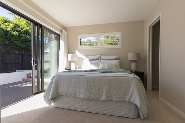 Bright, calm bedroom with a neatly made upholstered bed, layered white and pale blue bedding, matching bedside tables and lamps, beige walls and carpet, and large sliding glass doors opening to an outdoor terrace. A horizontal louvred window provides additional daylight, while simple white curtains and restrained furnishings create an airy contemporary feel.