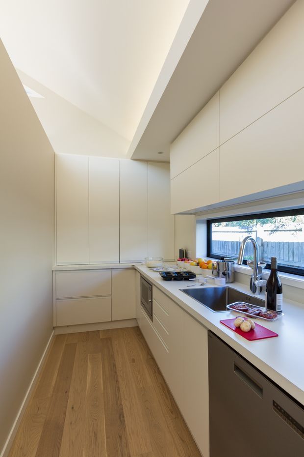 A narrow, contemporary galley kitchen with streamlined cream cabinetry, integrated appliances, a light engineered-stone benchtop, and a stainless-steel double sink beneath a wide horizontal window. Warm timber flooring contrasts with the pale cabinetry. A dramatic angled ceiling and recessed lighting create a bright, elongated space.