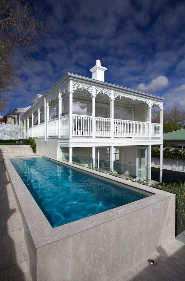 Exterior view of a substantial white two-storey house with an ornate wraparound verandah, decorative fretwork, turned balustrades, columns, and a prominent chimney. A long raised rectangular swimming pool occupies the foreground, with paved surrounds and glazed lower-level openings beneath the verandah. The image has a bright but dramatic outdoor atmosphere under a deep blue, cloud-filled sky.