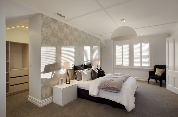 A bright, refined bedroom with a large upholstered bed, layered white and grey bedding, bedside tables and lamps, a black accent chair, plantation shutters, and patterned feature wallpaper. Built-in wardrobe or storage cabinetry is visible through the opening at left. The room has a carpeted floor, white decorative trims, and a suspended dome pendant light.