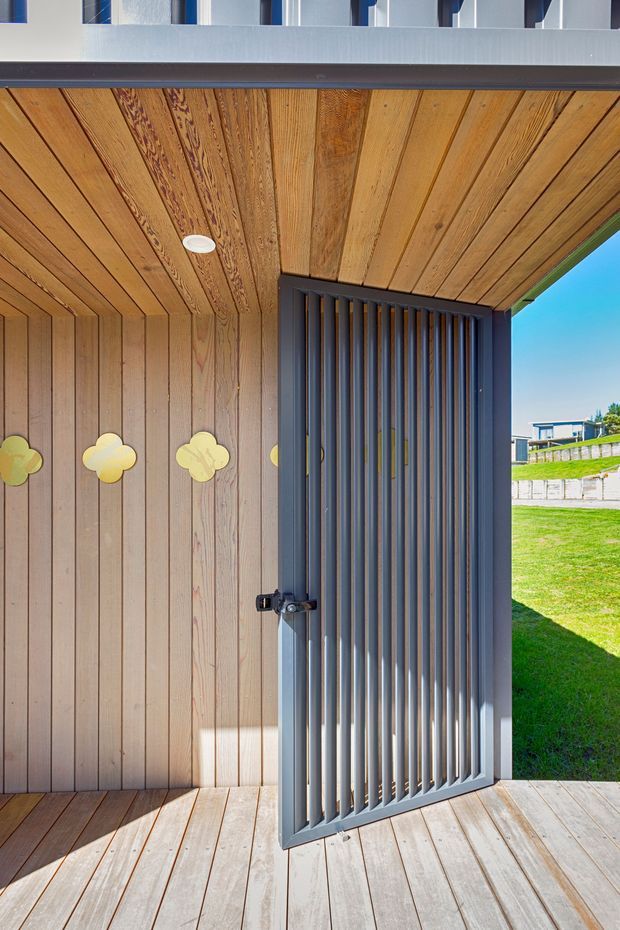 Close-up architectural view of a small outdoor entry or porch enclosure, featuring warm timber vertical wall cladding, a matching timber-lined soffit and decking, and a dark grey metal slatted security gate. Bright lawn and neighbouring buildings are visible beyond the open gate.