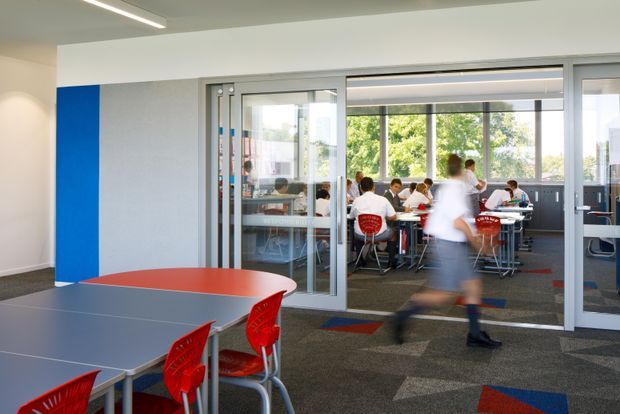 Bright contemporary school or institutional interior showing a flexible dining or common area in the foreground, with grey modular tables, red chairs, carpet tiles, and a vivid blue wall accent. Full-height glazed sliding or folding partitions open onto a busy secondary dining or collaborative space with additional tables and chairs. Linear recessed ceiling lighting and large windows create an airy, functional atmosphere.