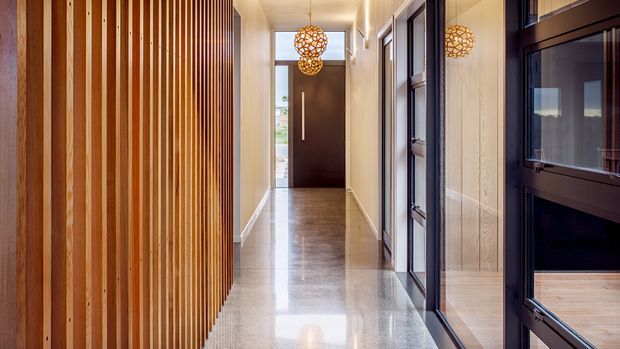 A contemporary residential hallway or entry corridor with polished concrete or terrazzo flooring, warm timber vertical slat screening along the left wall, pale textured walls, dark-framed internal glazing and doors on the right, and decorative spherical pendant lights leading toward a dark timber entry door. The space is bright, linear, refined, and warm, with reflections visible in the glossy floor and glazing.
