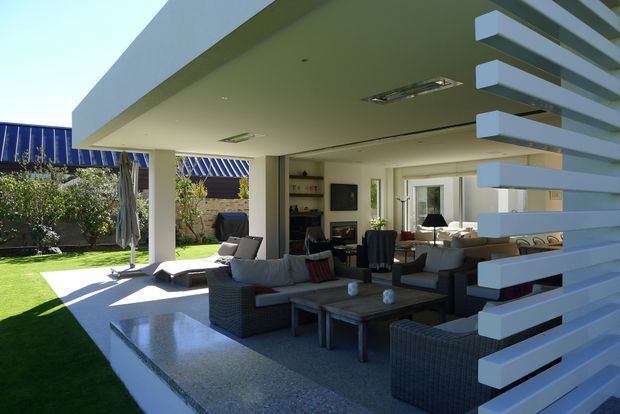 A contemporary covered outdoor living area adjoining a house, with a broad flat canopy, pale structural columns, polished light terrazzo-like flooring, and a horizontal white privacy screen. The space contains dark woven outdoor seating, a low coffee table, cushions, and a dining area visible beyond, opening onto a manicured lawn and garden.