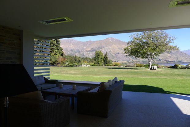 A shaded indoor-outdoor living area opens through a wide glazed opening onto an expansive lawn and landscaped grounds, with a mountain range and lake visible beyond. Dark wicker lounge furniture surrounds a low coffee table, while horizontal louvres screen one side of the opening. Recessed ceiling lights and a broad concrete or stone-toned floor create a modern, sheltered entertaining space.