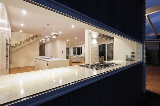 A contemporary open-plan kitchen viewed from an exterior or adjacent outdoor area through a large serving window. The kitchen features a long pale stone benchtop with integrated gas cooktop, white cabinetry, a tiled splashback and wall, pendant globe lights, recessed ceiling lighting, and timber flooring. A dining or living area is visible beyond the kitchen, with a stairway along the left side.