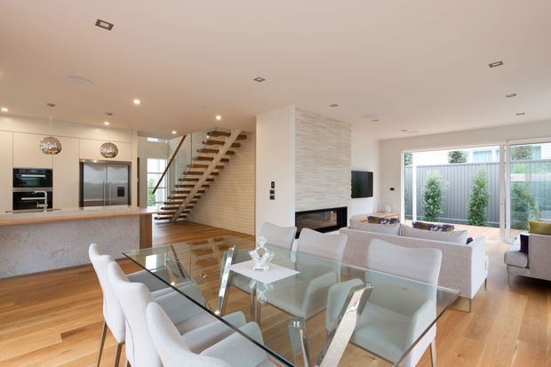 Bright open-plan contemporary interior combining a kitchen, dining area and living room. The space features warm timber flooring, a pale stone kitchen island, light cabinetry, a glass dining table with upholstered chairs, a white sectional sofa, recessed lighting, and a floating timber staircase beside a textured feature wall and fireplace. Large glazed doors open toward a planted outdoor area.
