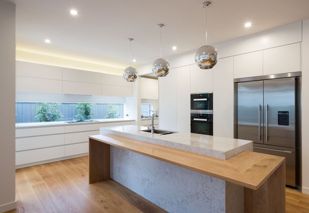 Bright contemporary kitchen with white handleless cabinetry, a large engineered-stone island with an integrated timber breakfast-bar surface, timber flooring, stainless-steel appliances, and three reflective spherical pendant lights. A long horizontal window provides natural light along the rear cabinetry.