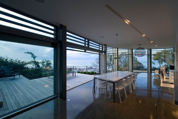 A contemporary open-plan dining and living space with extensive floor-to-ceiling glazing and louvred clerestory panels opening onto a timber deck. The room features a large light dining table, transparent dining chairs, polished dark flooring, recessed and linear track lighting, and views toward a coastal or waterfront landscape. The glazing and open connection create an airy indoor-outdoor setting.