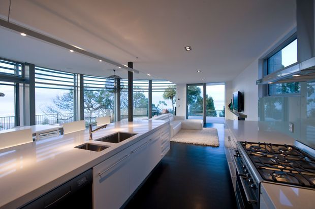 Open-plan contemporary kitchen and living area with a long white island, integrated sink, pale cabinetry, stainless-steel gas cooktop, and glossy light splashback. Dark timber flooring contrasts with the bright white surfaces. Floor-to-ceiling glazing and horizontal louvres provide expansive outdoor views and abundant natural light, while the living area extends beyond the kitchen.