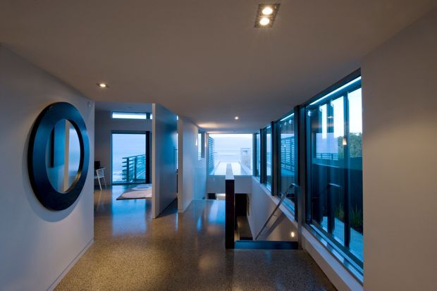 A contemporary interior hallway or landing with polished dark speckled flooring, white walls, recessed ceiling spotlights, and extensive floor-to-ceiling glazing along the right side. A large round black-framed mirror is mounted on the left wall, while an open stairwell and lower-level void appear in the centre-right. The corridor leads toward bright distant glazing and an outdoor or waterfront-like view, with blue evening light entering through the windows.
