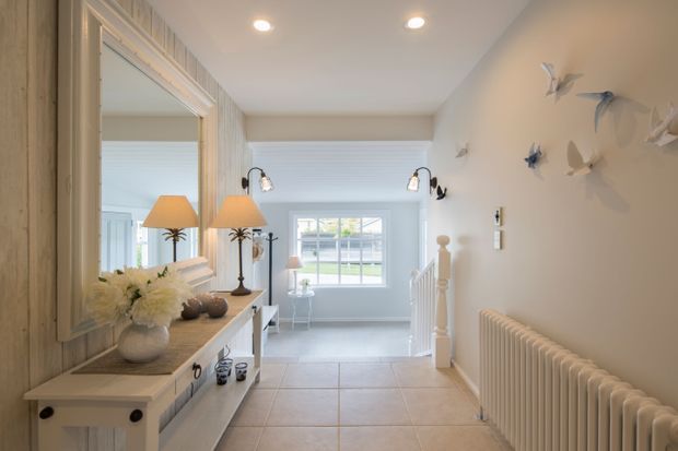 Bright, refined hallway or entry corridor with pale timber wall panelling, a large framed mirror, console cabinetry, decorative lamps, tiled flooring, a white radiator, and a stair opening. The corridor leads toward a light-filled sitting area with a large window; sculptural bird wall decorations add a coastal decorative character.