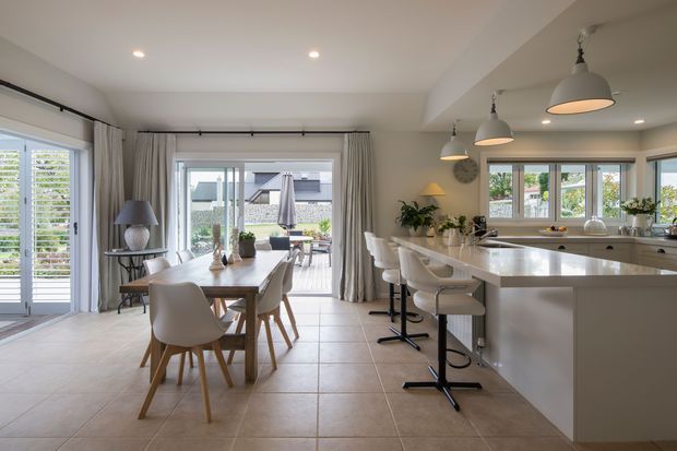Bright open-plan kitchen and dining area with a large white island, integrated seating, pendant lights, and a timber dining table. Floor-to-ceiling glazed doors and windows open toward an outdoor terrace, with neutral curtains, tiled flooring, and abundant natural light.