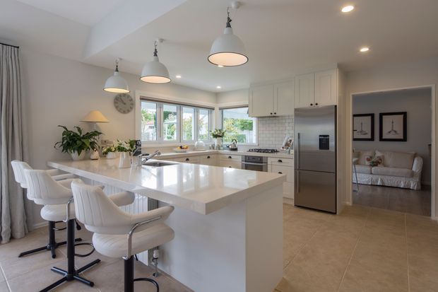 Bright, spacious open-plan kitchen with a large white island and four upholstered bar stools, white shaker-style cabinetry, a pale subway-tile splashback, stainless-steel appliances, and pendant lighting. A dining/living area is visible beyond, with a neutral palette, tiled floor, and large corner windows providing abundant natural light.