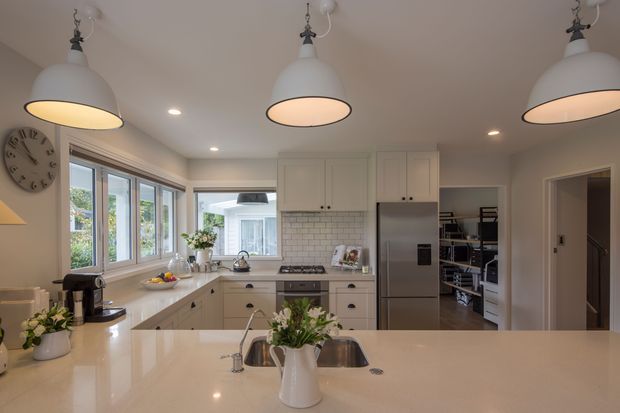Bright, spacious contemporary kitchen with a large pale stone island, white cabinetry, stainless-steel appliances, subway-tile splashback, and pendant lighting. The room includes a broad bank of windows, a walk-in pantry visible beyond the refrigerator, and an adjoining hallway. Neutral finishes and floral accents create a clean, inviting atmosphere.