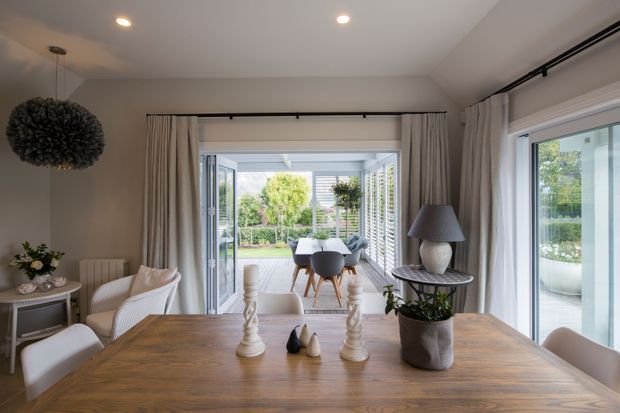 Bright contemporary dining room centred on a large natural timber dining table, with upholstered dining chairs, decorative pendant lighting, and neutral curtains. Wide glazed doors open to a covered outdoor dining or patio area, creating a strong indoor-outdoor connection. The room has recessed ceiling lights, light neutral walls, and a restrained, refined palette.