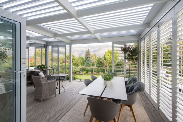 Bright contemporary enclosed outdoor room or sunroom with a dining area and separate lounge seating. The space features grey timber-look decking, a large outdoor dining table, woven armchairs, extensive white horizontal louvre screens, a glazed door, and a translucent slatted roof supported by white structural beams. The open sides look onto a landscaped garden and distant mountain views.