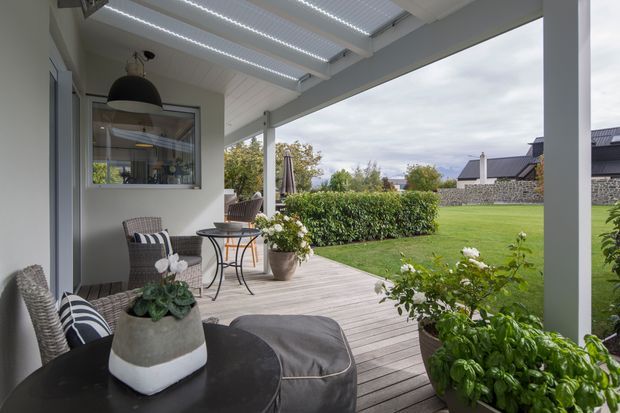 Covered outdoor veranda with timber decking, outdoor seating, potted plants, and a dining area overlooking a manicured lawn and neighbouring homes. The space has white structural posts, a translucent corrugated roof, and a relaxed contemporary outdoor-living character.