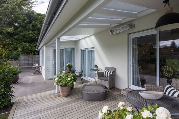 A covered outdoor deck and patio adjoining a light-coloured single-storey home. The space features timber decking, sliding glass doors, a white pergola-style roof with translucent slats, outdoor wicker seating, potted plants, and a paved garden walkway. The setting feels bright, leafy, and residential.