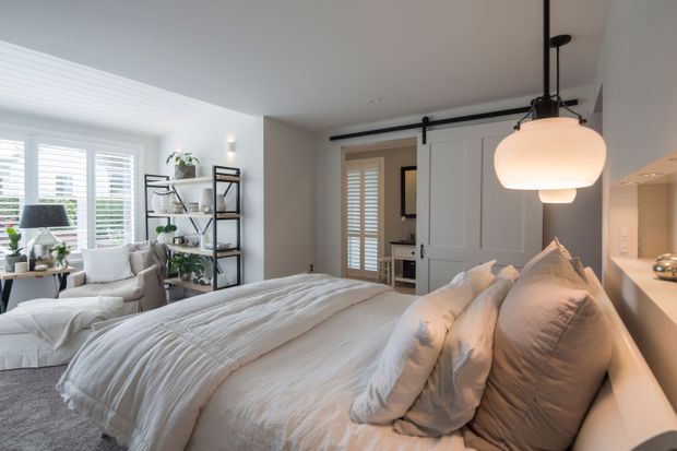 Bright, softly styled bedroom with a large upholstered bed dressed in layered white bedding, abundant pillows, and a neutral grey rug. The room includes a white lounge chair by shuttered windows, open black metal shelving with plants and decor, white paneled cabinetry, and a striking oversized pendant light suspended from a black ceiling rail. A shuttered doorway leads to an adjacent space.