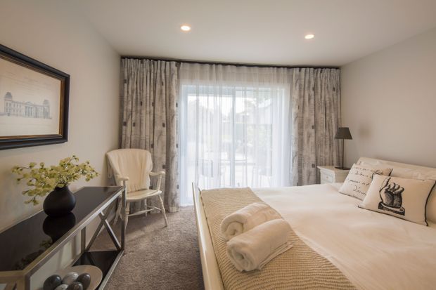 A refined bedroom with a large bed dressed in cream and beige linens, textured throw, rolled towels, and decorative cushions. Floor-to-ceiling sheer curtains and patterned drapes frame a glazed door or window, while a pale upholstered armchair, dark console table, framed artwork, bedside furniture, and recessed ceiling lights complete the room. The palette is warm, soft, and neutral with a comfortable hotel-like atmosphere.