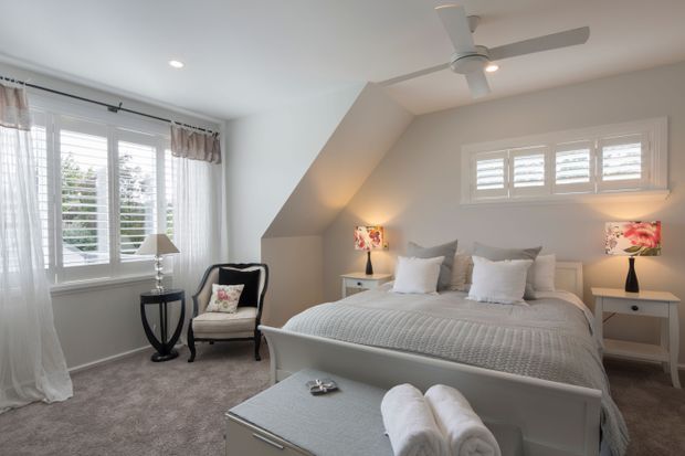 Bright, elegant bedroom with a large upholstered bed, white bedding, matching bedside tables and floral lamps. The room features a sloped ceiling, ceiling fan, plantation shutters, sheer curtains, grey carpet, and a upholstered storage bench at the foot of the bed. The palette is predominantly soft white, light grey, and muted taupe with small floral accents.