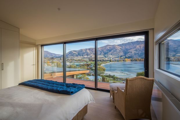 A bright contemporary bedroom with a large bed, built-in light cabinetry, timber flooring, and full-height corner glazing overlooking a lake and mountainous landscape. A woven armchair sits beside the windows, while the room has a calm, restrained palette of white, beige, and natural timber tones.
