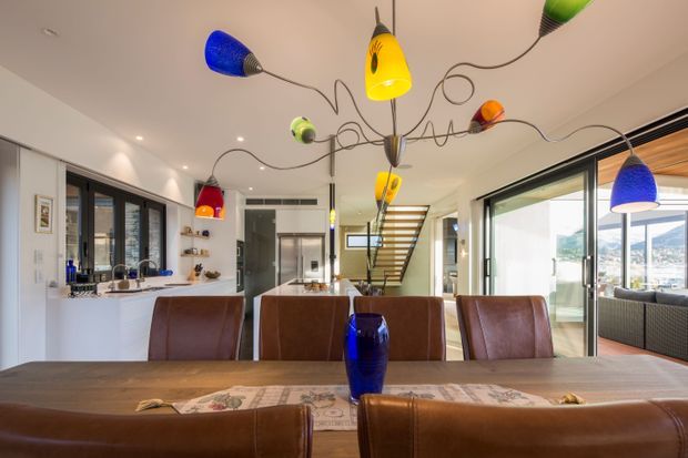 Bright open-plan dining and kitchen interior with a large timber dining table, brown leather dining chairs, white kitchen cabinetry, extensive glazing, and a colourful sculptural pendant light. A stairway and outdoor views are visible beyond the dining area.