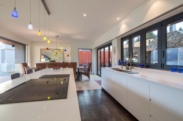 Bright contemporary open-plan kitchen and dining area with a long white island, integrated black induction cooktop, extensive white cabinetry, dark timber flooring, and black-framed windows and doors. A dining table and colourful pendant lights extend through the open space, while a stone-clad exterior wall is visible beyond the glazing.