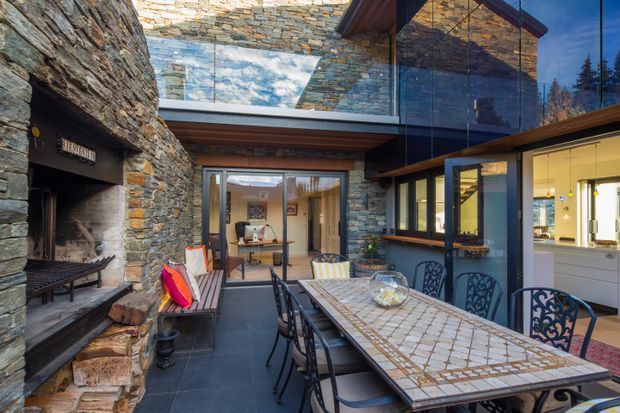 A sheltered courtyard dining area beside a large rough-stone fireplace, with a long tiled dining table and metal chairs. Stone-clad walls, dark timber beams, extensive glazing, and a skylight create a rustic yet refined indoor-outdoor setting. A kitchen is visible through the glazing on the right, while a seating area appears beyond the central doors.