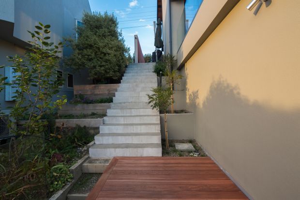 A landscaped external walkway between modern residential buildings, featuring a broad flight of pale concrete steps leading upward from a warm timber deck. Planted garden beds, small trees, rendered walls, metal-framed glazing, and contemporary exterior lighting define the scene.