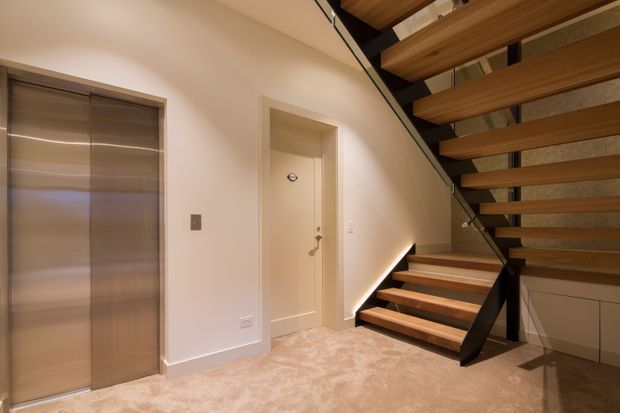 Interior view of a contemporary stair hall with an open-riser timber staircase, dark structural supports, integrated under-stair cabinetry, a carpeted floor, and a flush metallic lift door beside a plain internal door. Warm concealed stair lighting accentuates the clean-lined detailing.